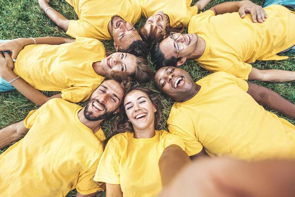 公園で草の上に横たわる自撮り写真を撮る幸せな多文化の友人 大学キャンパスの多様な学生 — ストック写真