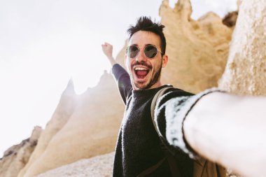 Sırt çantasıyla dağda yürüyüş yapan mutlu genç adam akıllı telefon ve kamera ile selfie çekiyor. Seyahat macerası konsepti.