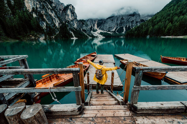 Rear view portrait of a hiker with arms outstretched visiting alpine lake at Braies, Italy. Happy hiker wearing yellow jacket and backpack enjoy the nature landscape at autumn. Wanderlust and travel.