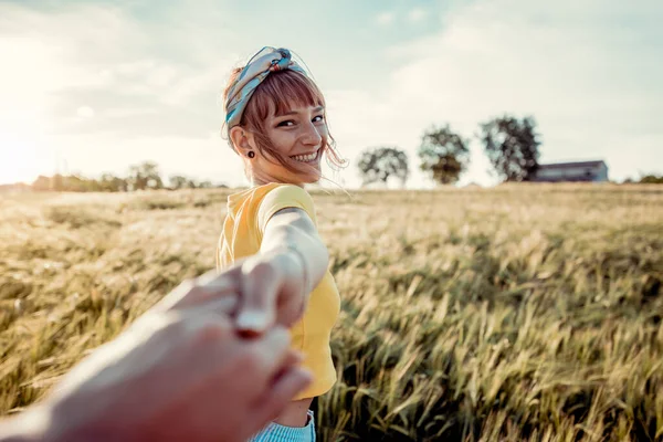 日没時に小麦畑を歩きながらボーイフレンドの手を握っている幸せな若い女性の肖像画 自然の中で旅行を楽しむカップル ストックフォト