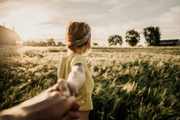 日没時に小麦畑を歩きながらボーイフレンドの手を握っている幸せな若い女性の肖像画 自然の中で旅行を楽しむカップル ロイヤリティフリーのストック画像