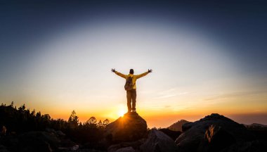 Erkek yürüyüşçü dağa tırmanıyor - güçlü yürüyüşçü elleri uçurumun tepesinde duruyor günbatımının tadını çıkarıyor