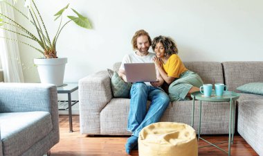 Evdeki kanepede oturup bilgisayar izleyen çok ırklı genç bir çift mutlu karı-koca bilgisayar hizmetleri kullanıyor teknoloji yaşam tarzı konsepti. 
