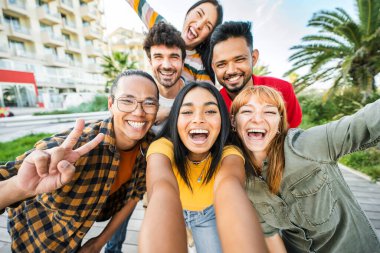 Çok ırklı arkadaşlar dışarıda akıllı cep telefonlarıyla selfie çekiyorlar mutlu gençler şehir sokaklarında konuşarak eğleniyorlar güneşli bir günde kızlar ve erkeklerle takılıyorlar. 