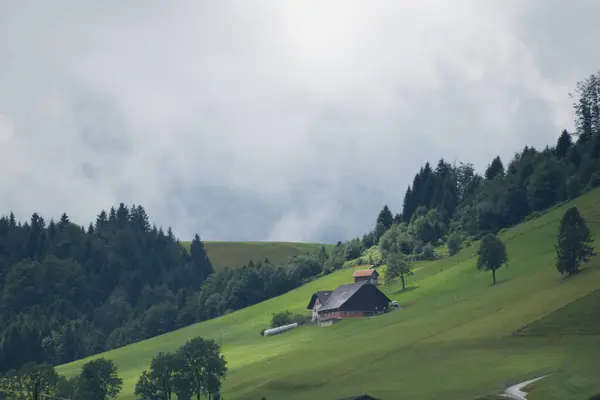 A solitary farmhouse sits amidst lush alpine hills, partially veiled by clouds, in the tranquil Swiss countryside.