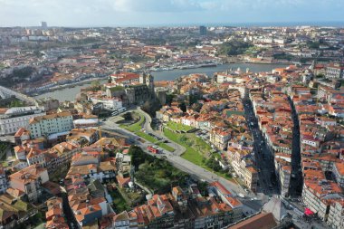 Sunlight bathes the historic district of Porto, illuminating its colorful buildings and the intricate web of streets. This aerial image showcases the city's architectural beauty and urban layout, with a clear view leading to the Douro River. The warm