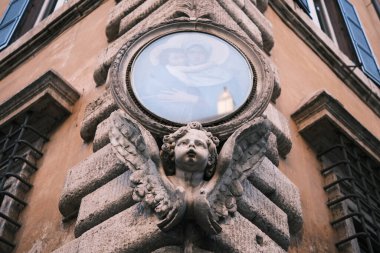 An angelic figure cradles a round window, its cherubic face turned upwards, embodying the artistic spirit that adorns the historical buildings of Rome, a fusion of divine inspiration and architectural elegance in Italy's capital.