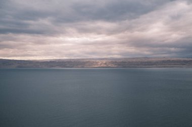 The tranquil waters of the Dead Sea lie beneath a canopy of moody skies in Jordan, creating a serene and contemplative landscape. The calm expanse of water juxtaposed with the dramatic weather conveys a sense of timelessness and natural beauty.