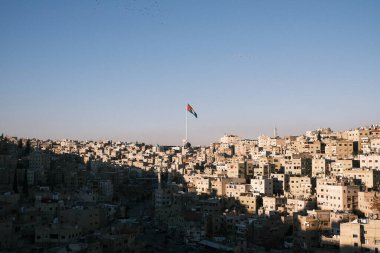 A majestic Jordanian flag waves over the sprawling cityscape of Amman at dusk. This image perfectly encapsulates the vibrant life and soul of Jordan's capital. The warm glow of the sunset bathes the city in soft light, highlighting the dense architec