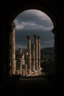 This dramatic view through an archway frames the ancient columns of a historical site in Jordan, offering a glimpse into the past. The architectural grandeur of the scene is accentuated by the contrast of light and shadow, inviting contemplation of t