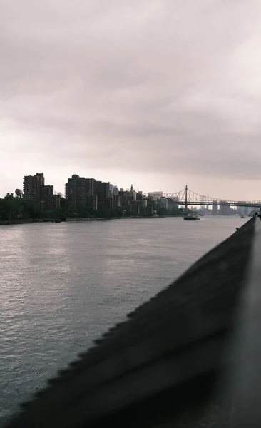 The moody overcast sky sets a dramatic backdrop for the Queensboro Bridge and the silhouette of New York City's skyline, capturing the city's dynamic atmosphere.