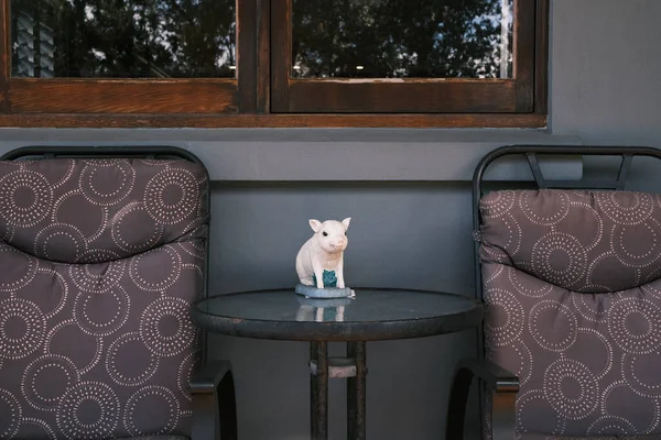 This charming image features a ceramic pig figurine poised on a round table between two patterned chairs on a cozy porch. The scene is a delightful representation of home decor and the personal touches that make a space feel welcoming. It's a snapsho