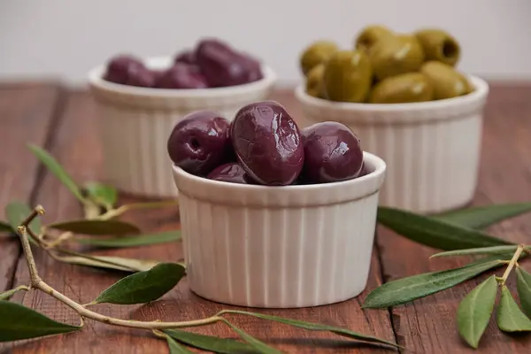 Yuvarlak kaselerde siyah ve yeşil zeytinler, zeytin yapraklı, ahşap bir arka planda.
