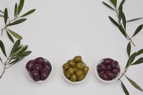 Yuvarlak kaselerde siyah ve yeşil zeytinler, beyaz bir zemin üzerinde zeytin yaprakları ile metin için boşluk ile