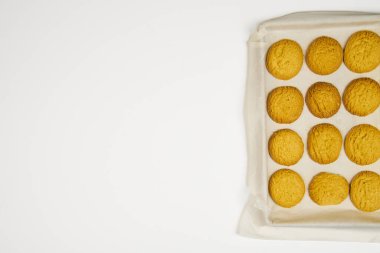 Bir tepsi taze pişmiş kurabiye kağıdın üzerinde beyaz bir zeminde duruyor. Kurabiyeler küçük ve yuvarlak, hepsi aynı boyutta. Tepsi alüminyumdan yapılmış ve kurabiyeler sıra sıra dizilmiş..