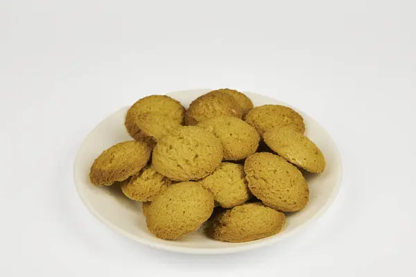 Beyaz arka planda bir tabak kurabiye duruyor. Kurabiyeler küçük ve yuvarlak ve hepsi aynı renk. Tabak boş ve kurabiyeler düzgün bir yığın içinde.