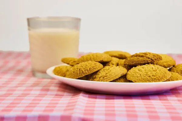 Bir tabak kurabiye pembe ve beyaz kareli masa örtüsünün üzerinde bir bardak sütün yanında duruyor. Kurabiyeler piramit şeklinde düzenlenir ve en büyük kurabiye en üstte olur. Konfor ve zevk kavramı.