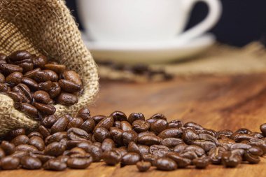 Kavrulmuş kahve çekirdekleri ile kavrulmuş bir jüt torba, yanında da bir fincan taze kahve.