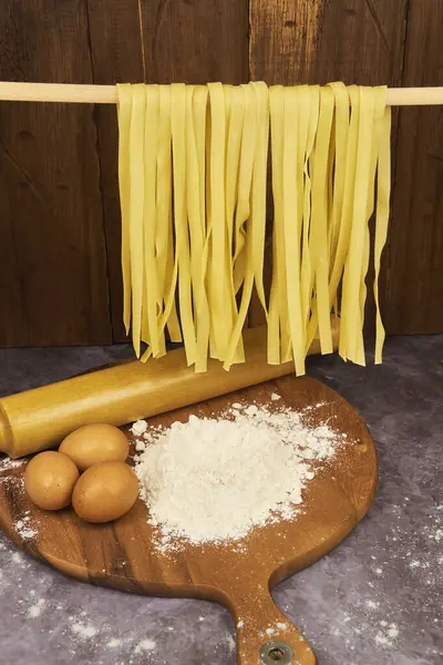 Taze yumurta fettuccine eriştesi, ön planda kurutuluyor, un ve yumurta içeren ahşap bir masanın üzerinde asılı duruyor, hazırlık için temel malzemeler..