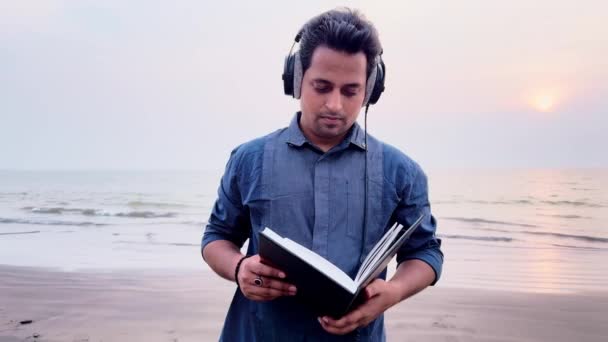 Człowiek Słuchawkami Czytający Książkę Spacerujący Spokojnej Plaży Zmierzchu — Wideo stockowe