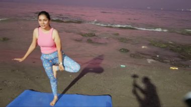 Alacakaranlıkta plajda yoga yapan bir kadın okyanus zeminli mavi bir paspasın üzerinde ağaç pozu veriyor.