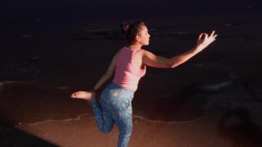 Aktif genç bir kadın, denge ve sükunet üzerine odaklanarak, alacakaranlıkta bir plajda yoga pozu veriyor.