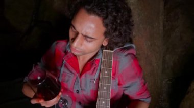 Şarap kadehi ve akustik gitarla konuşan sarhoş bir adam loş bir ortamda oturmuş karamsar ve romantik bir ortam yaratıyor.