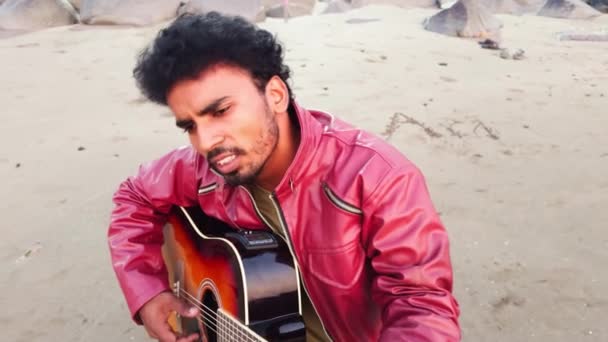 穿着红色夹克的男人在沙滩上弹奏着声吉他 背景音乐为岩石伴奏 — 图库视频影像