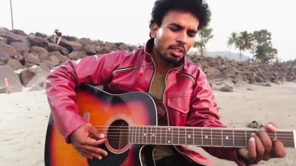 穿着红色夹克的男人在沙滩上弹奏着声吉他 背景音乐为岩石伴奏 — 图库视频影像