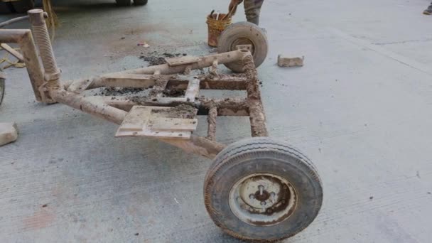 建設現場のコンクリート地面に車輪が付いている古い車の錆ついた金属フレームを動かす労働者 — ストック動画
