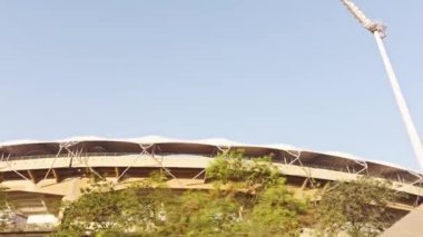 Modern bir stadyumun dış görüntüsü geniş bir direğe sahip açık bir gökyüzü etrafında yeşil yapraklar ile çevrili bu sırada araba Asya, Hindistan otoyolunda ilerliyor. 