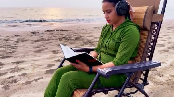 バックグラウンドで岩と砂でリクライニングチェアに座ってヘッドフォンを着用しながらビーチで本を読む集中女性 — ストック動画