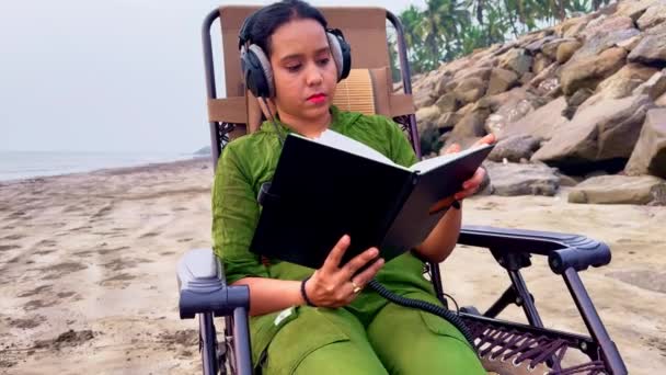 Mujer Enfocada Leyendo Libro Playa Mientras Usa Auriculares Sentados Una — Vídeos de Stock
