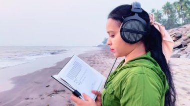 Sahilde, deniz kenarında kulaklıkla müzik dinlerken kitap okuyan odaklanmış bir kadın.