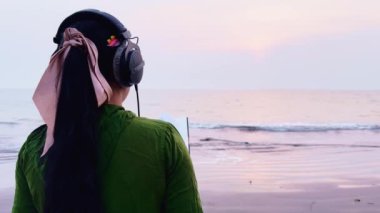 Sahilde, deniz kenarında kulaklıkla müzik dinlerken kitap okuyan odaklanmış bir kadın.
