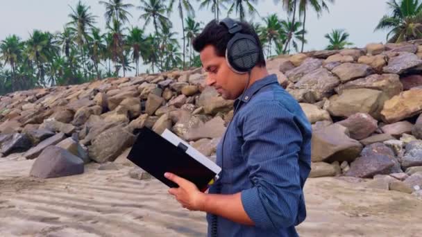 海でヘッドフォンで音楽を聞いている間 本を読んで歩いている男 — ストック動画