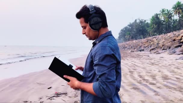 Deniz Kenarındaki Kulaklıklarla Müzik Dinlerken Kitap Okuyarak Yürüyen Adam — Stok video