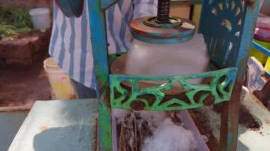 Arka planda renkli şurup şişesi olan bir sokak standında buz kesme makinesiyle buz külahı yapan kişi.