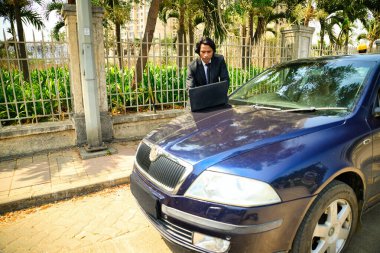 Kaputta dizüstü bilgisayarda çalışan bir iş adamı, sokakta park etmiş mavi bir araba, arka planda çit ve yeşillik olan genç bir avukat, Hintli, Online çağrı, yolda buluşmalar, 30 'lu yaşlarda bir adam..