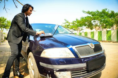 Araba kaputunda dizüstü bilgisayarda çalışan mavi bir arabanın kaputuna yerleştirilmiş kaldırıma park etmiş bir avukat. Arka planda bir çit, yeşillik, açık hava işi yan yolda bir girişimci, Hintli, 30 'lu yaşlarda..