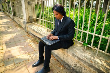 Dışarıda dizüstü bilgisayarda çalışan bir iş adamı, taştan bir çıkıntının üzerinde otururken, yeşil bitki arka planına sahip metal çitlerin yanında, odaklı genç bir iş adamı, cadde yolunda oturuyor, Hintli..