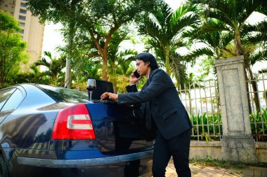 İş adamı dizüstü bilgisayarda ve telefonla çalışıyor takım elbiseli bir adam, bir arabanın bagajına yaslanıyor ve bir bilgisayar kullanıyor. Araba bir caddeye park edilmiş, ağaçlar ve çitler var, Hintli, 30 'larında..