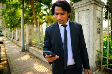 Yürüyen ve akıllı telefon kullanan bir iş adamı siyah takım elbiseli ve kravatlı bir adam cep telefonuna bakarken kaldırımda yürür. Açık havada, ağaçlar ve çitlerle sosyal medyayı kontrol ediyor, Hintli, odaklanmış..