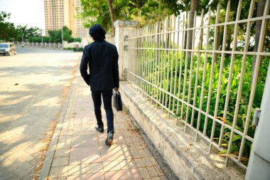 Siyah takım elbiseli bir adam çantasıyla kaldırımda yürüyor, çitin yanında bir laptop çantası taşıyor, iş adamının arkası görünüyor, sokakta yürüyor, ağaçlar ve binalar arka planda, ofise giden çalışan, Hintli bir çalışan..