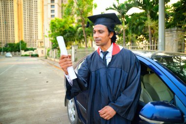 Diplomasını arabasının yanında tutan mezuniyet elbisesi ve şapkası giyen bir adam mutlu görünüyor, mutlu görünüyor, sertifikaya bakıyor, ağaçlar var, Hintli..