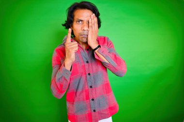 Bir gözünü kapayan ve tehdit eden bir adam, birini tehdit eden bir adam, tokat yiyen kızgın bir adam, yeşil bir arkaplan karşısında duran kareli gömlek, işaret parmağını kaldırırken, Hintli, yüzüne tokat atan, 30 'lu yaşlarda,.