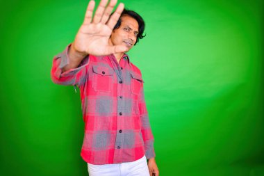 Man in checkered shirt gesturing stop, Smiling men end gesture, Looking at camera, And white pant stands, in front of a green background, Holding up his hand towards the camera, Indian, Gestured, 30s. clipart