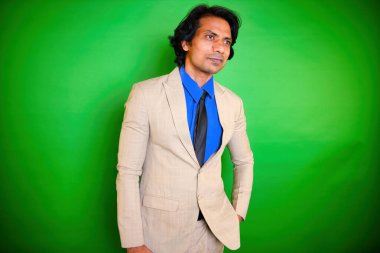 Gülümseyen otel müdürü bej takım elbiseli, poz veren, genç bir iş adamı bakışları kaçıran bir girişimci, Hintli, mavi gömlekli ve siyah kravatlı, yeşil arka planda duran, 30 'lu yaşlarda, Asyalı bir adam..