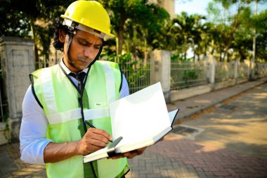 Caddede kitap yazan bir mühendis, inşaat işçisi yolda panoya yazı yazıyor, sarı bir şapka takıyor, yolda duruyor. Ağaçlar çitle çevrili, odaklanmış ustabaşı rampayla duruyor, alçak açı, Hintli..