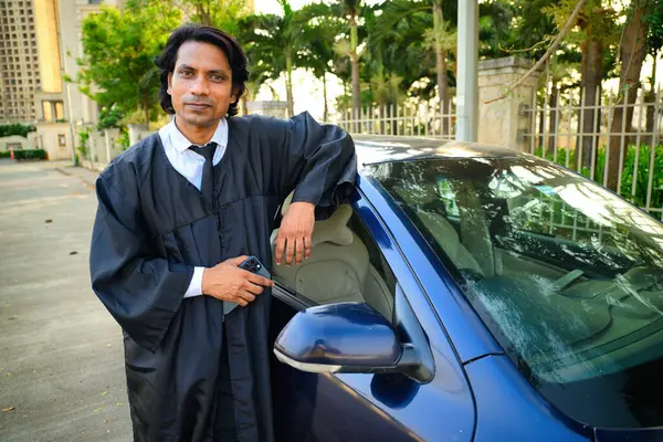 stock image Lawyer standing next to a blue car outdoors, A man in lawsuit standing next to a blue car, holding a smart phone, Young judge, Outside setting, trees and buildings, Indian, smiling, Looking at camera.